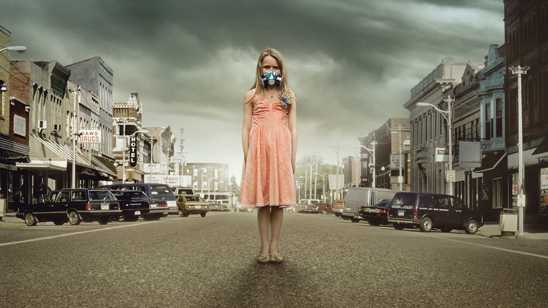HD desktop wallpaper from the movie The Crazies showing a young girl in a pink dress wearing a gas mask, standing alone on a deserted town street under a cloudy sky.