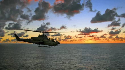HD PC wallpaper featuring a Bell AH-1 SuperCobra military helicopter flying over the ocean at sunset with dramatic clouds in the sky.