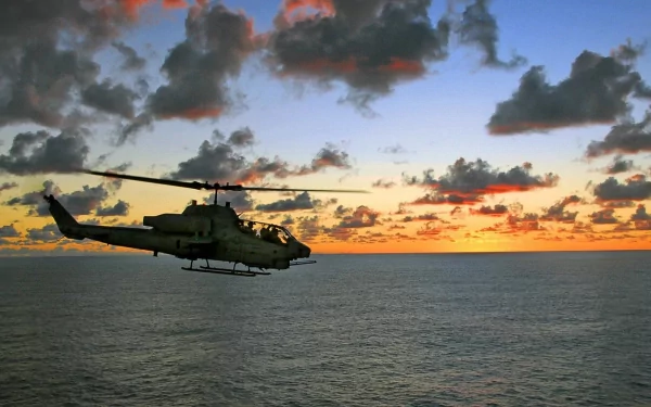 HD PC wallpaper featuring a Bell AH-1 SuperCobra military helicopter flying over the ocean at sunset with dramatic clouds in the sky.