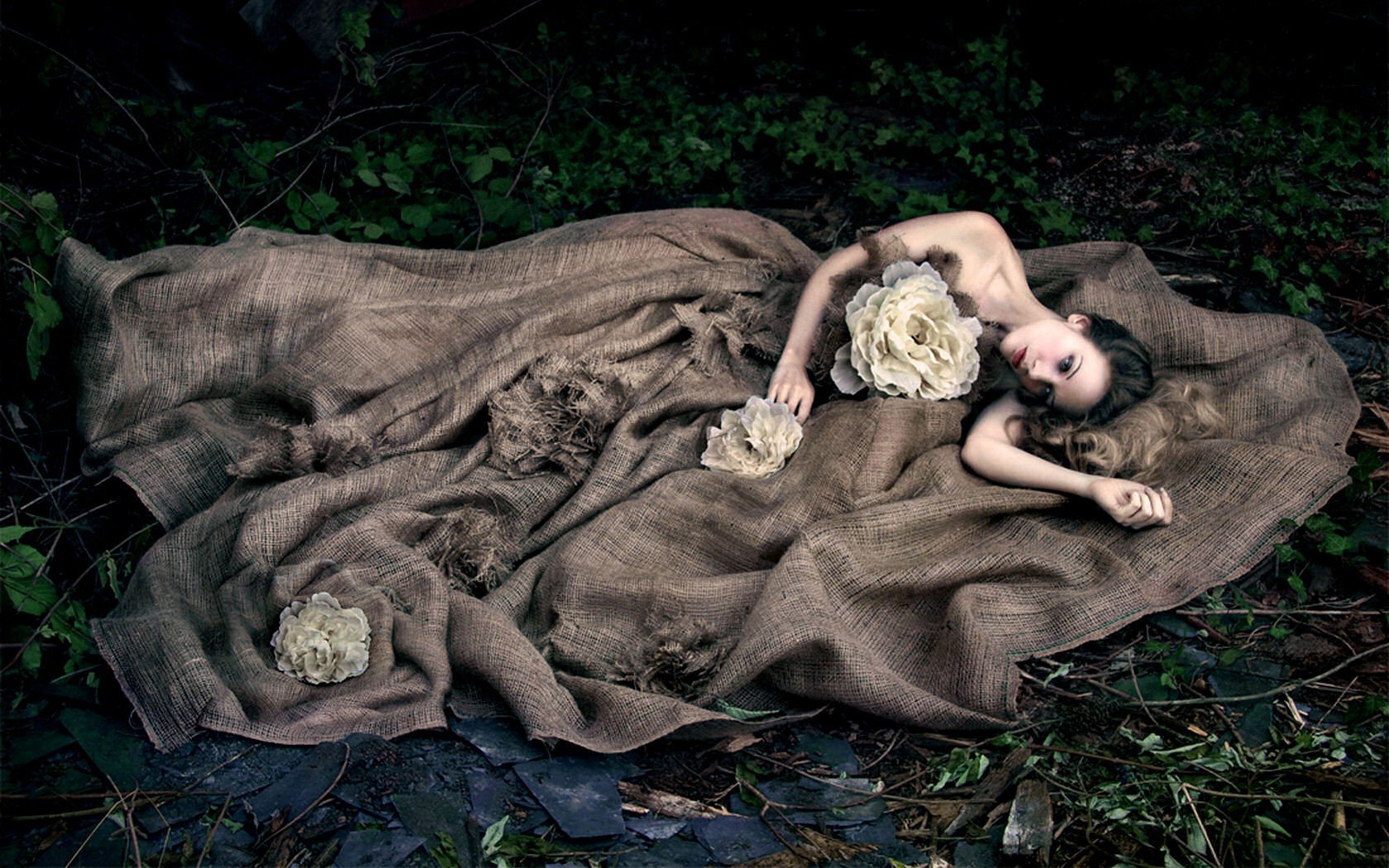 HD PC desktop wallpaper featuring a woman lying on a textured brown fabric in a natural, forest-like setting, holding large cream-colored flowers.