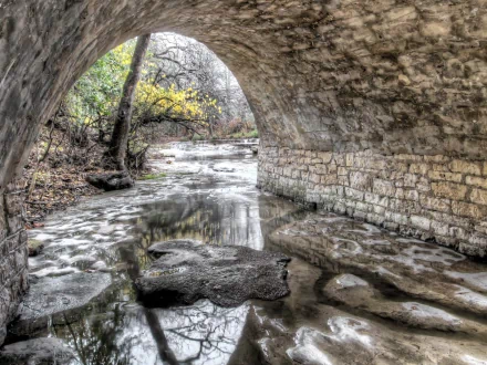 HD desktop wallpaper featuring a serene natural scene viewed through a stone man-made tunnel, with a calm river flowing beneath and trees in the background.