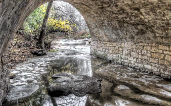 HD desktop wallpaper featuring a serene natural scene viewed through a stone man-made tunnel, with a calm river flowing beneath and trees in the background.