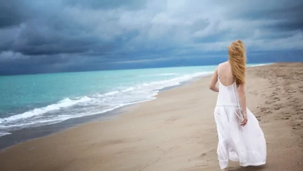A woman in a white dress stands on a beach, facing the ocean under a cloudy sky; HD desktop wallpaper and background.
