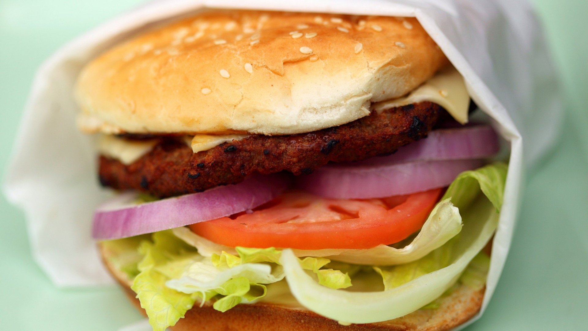 HD PC desktop wallpaper featuring a close-up of a fresh burger with lettuce, tomato, red onion, cheese, and a sesame seed bun.