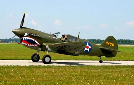 HD desktop wallpaper showing a vintage military aircraft with shark mouth nose art, parked on a runway with clear skies in the background.