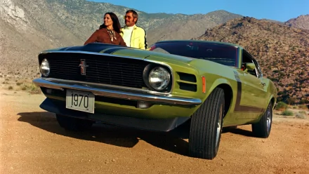 A green 1970 Ford Mustang Boss 302 muscle car parked on a desert road with two people sitting on its hood against a mountainous backdrop.