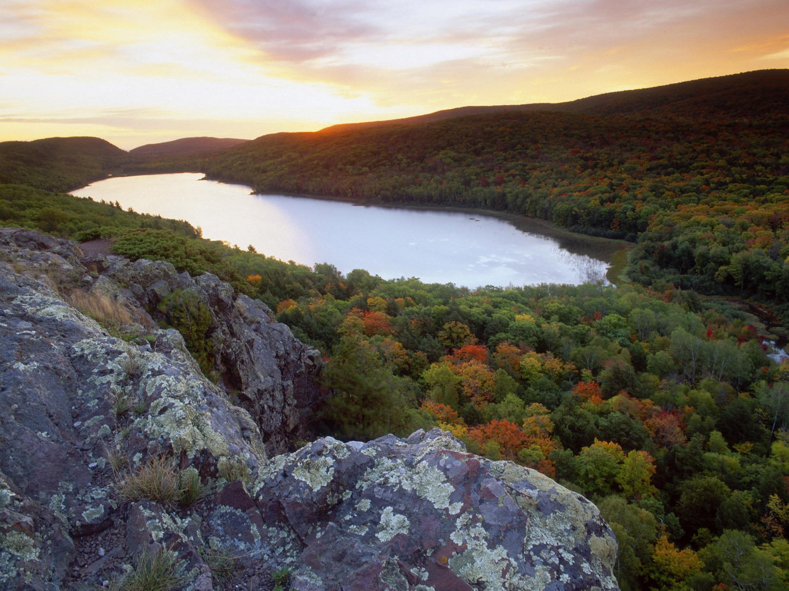 HD desktop wallpaper of a forest and lake, capturing a serene nature scene at sunset. The background features rugged cliffs and vibrant autumn foliage surrounding a tranquil body of water.