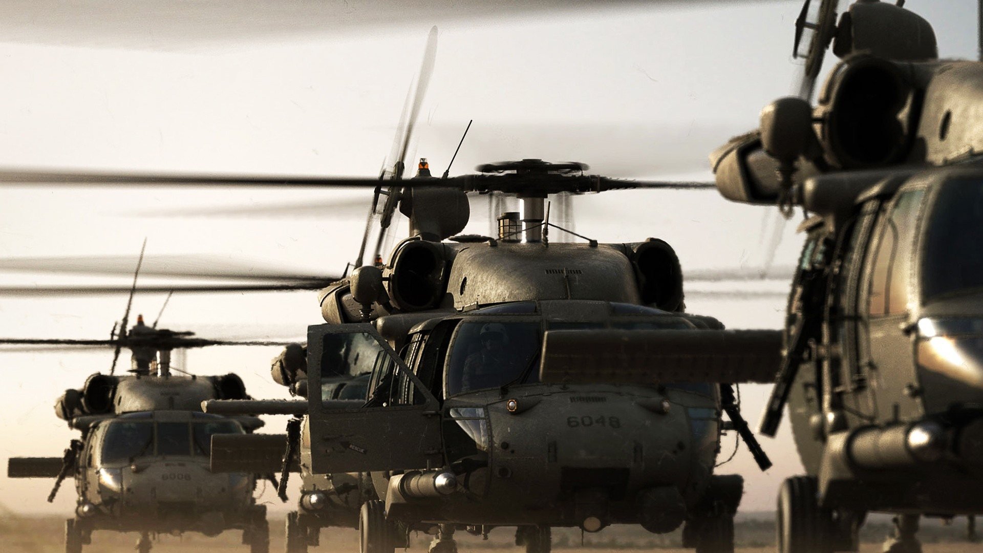 HD PC desktop wallpaper background showing military Sikorsky HH-60 Pave Hawk helicopters in formation on a dusty airfield, rotors spinning in low light.