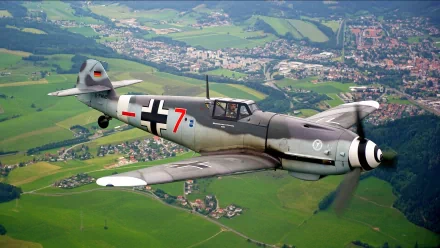 HD desktop wallpaper featuring a military Messerschmitt Bf 109 aircraft flying over a green landscape with a town in the background.