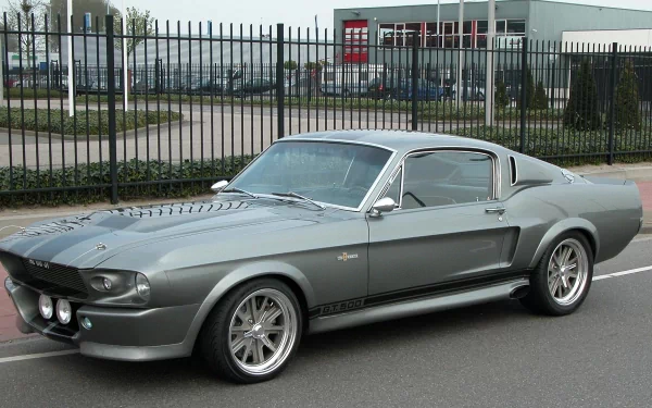HD PC desktop wallpaper featuring a classic silver Ford Mustang GT500 parked on an urban street with a metal fence and buildings in the background.