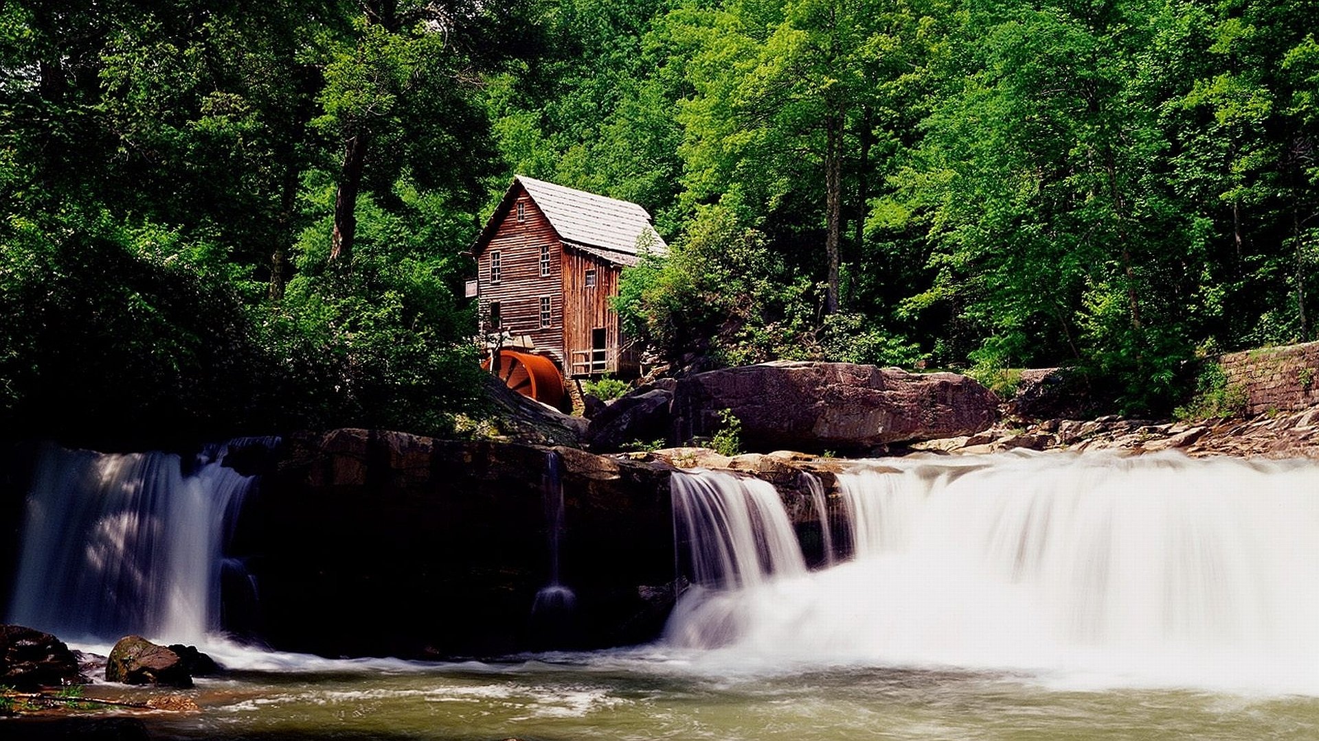 HD PC desktop wallpaper featuring a man-made watermill beside a flowing waterfall, surrounded by lush green trees and natural rock formations.
