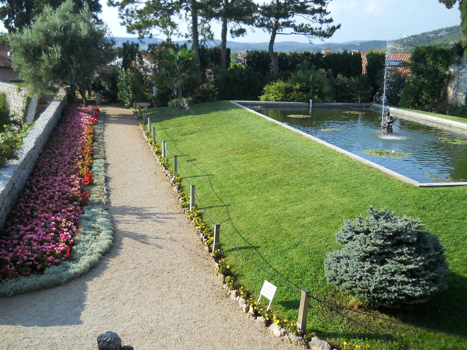 Photography of a manicured park with a gravel path, colorful flowerbeds, trimmed lawn and a rectangular reflecting pool with fountain; 2K Quad HD PC desktop wallpaper and background.