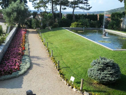 Photography of a manicured park with a gravel path, colorful flowerbeds, trimmed lawn and a rectangular reflecting pool with fountain; 2K Quad HD PC desktop wallpaper and background.
