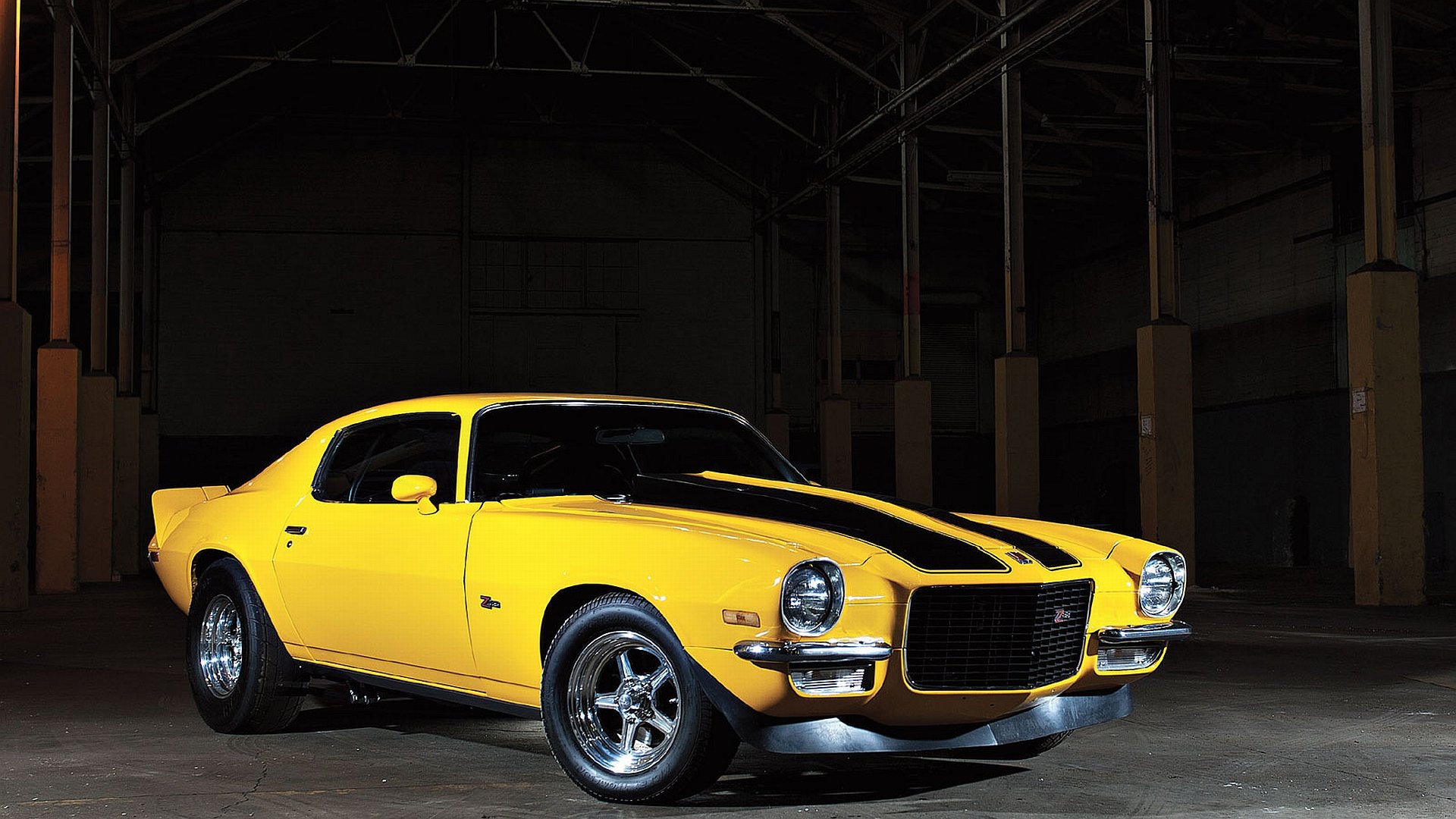 HD PC desktop wallpaper and background: yellow Chevrolet muscle vehicle with black racing stripes parked in a dim industrial garage.