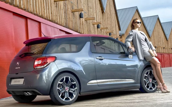 A stylish woman leans against a sleek gray car, showcasing a modern and chic vibe, with a backdrop of wooden structures, creating a captivating HD desktop wallpaper.