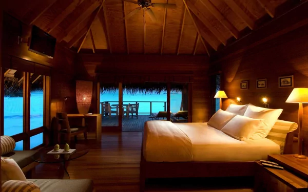 A warm, man-made room with wooden interiors and a large bed, opening to a balcony overlooking a serene blue ocean, captured in HD for a PC desktop wallpaper.
