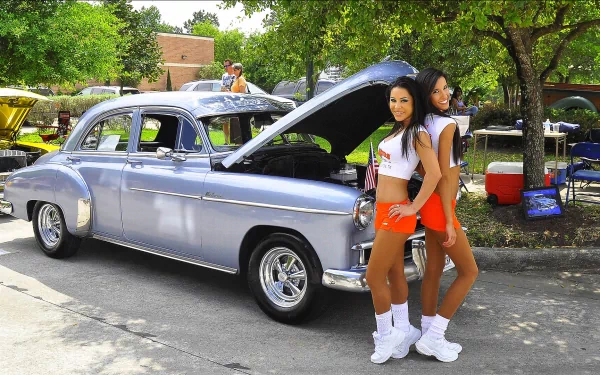 HD PC desktop wallpaper featuring two women posing next to a classic silver car at an outdoor car event under green trees.