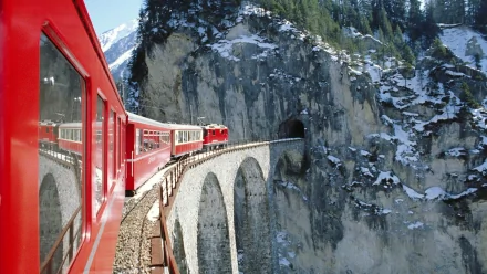 HD PC desktop wallpaper featuring a red RhB train crossing a stone arch bridge over a deep mountainous gorge with snowy cliffs in the background.