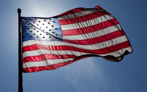 HD desktop wallpaper featuring an American flag waving gracefully against a clear blue sky with the sun creating a silhouette effect behind the flag.