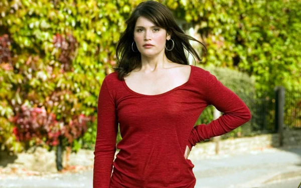Gemma Arterton stands confidently outdoors, wearing a red top, with lush greenery in the background. This HD image serves as a striking desktop wallpaper.
