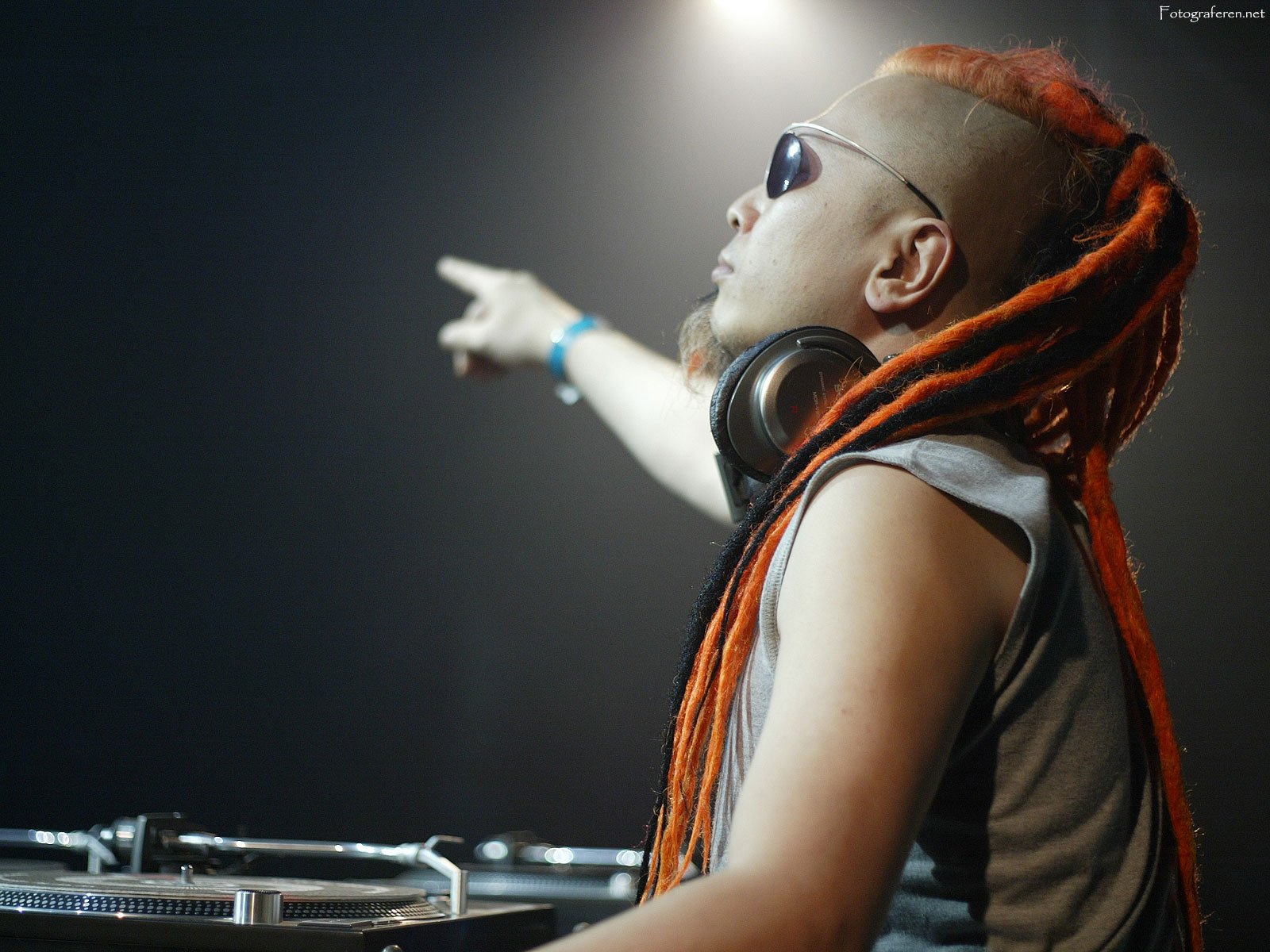 DJ with orange dreadlocks and sunglasses pointing into the distance while wearing headphones, set against a dark background. High-definition desktop wallpaper.