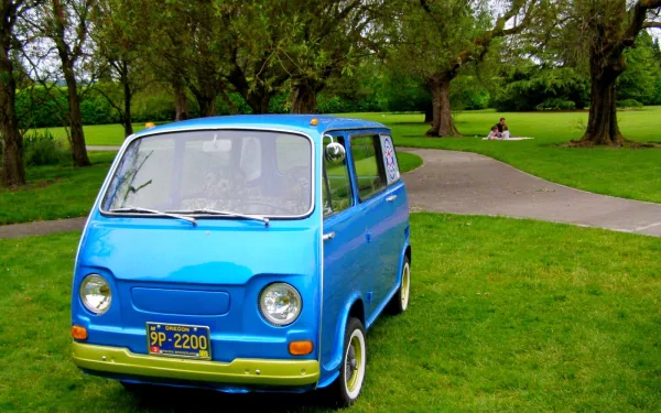 Blue vintage Subaru microvan parked on a grassy park lawn beside a winding path, trees in the background — HD PC desktop wallpaper and background.