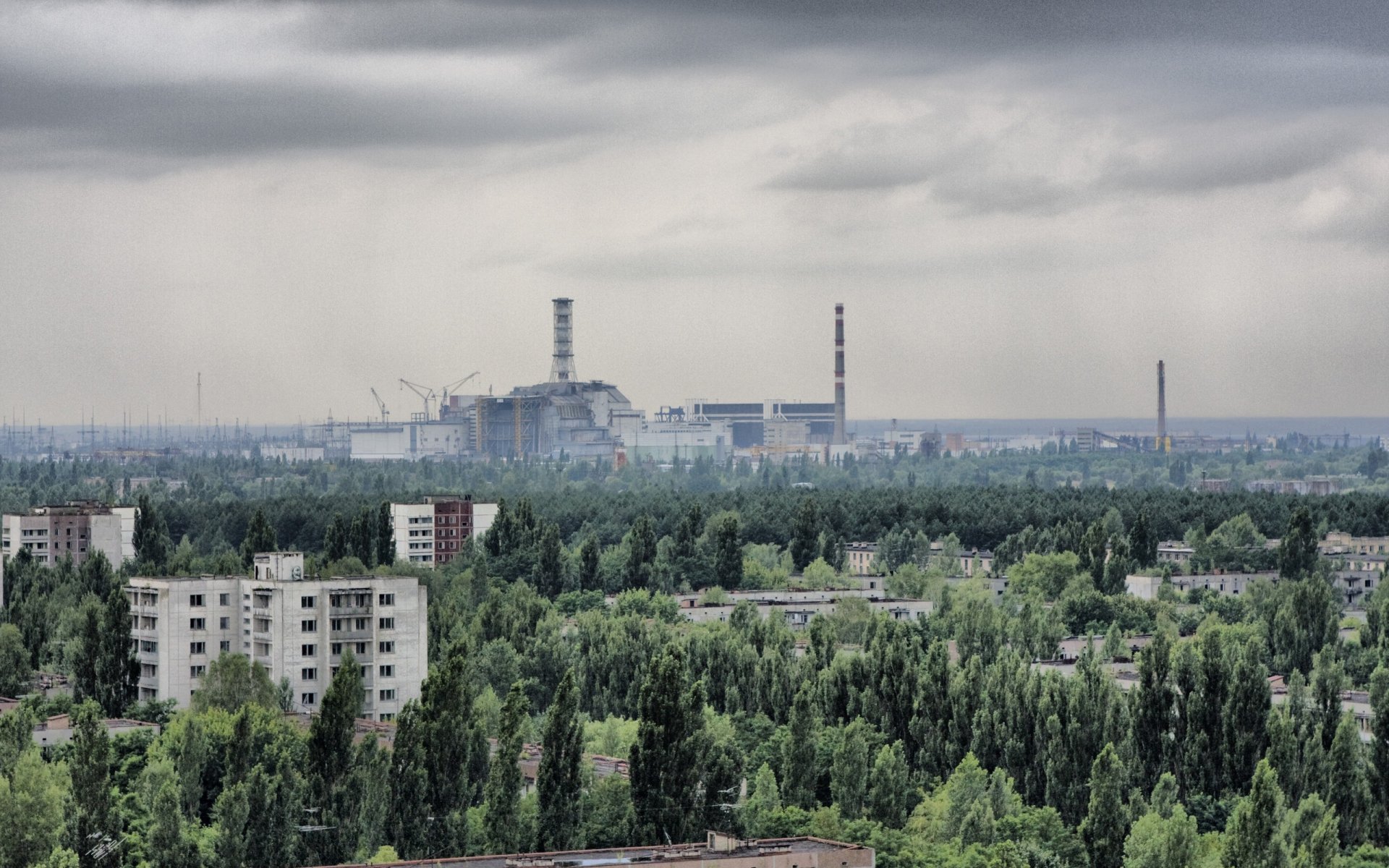 Pripyat, Chernobyl skyline: abandoned apartment blocks and overgrown trees, distant reactor and factory smokestacks beneath a brooding gray sky — 2K Quad HD desktop wallpaper