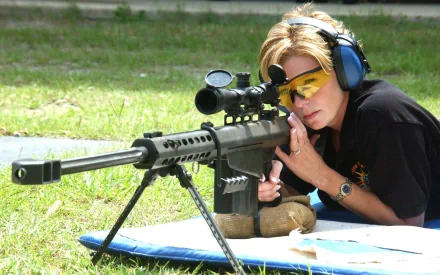 A woman expertly aims a rifle on a grassy range, wearing protective gear. This striking HD wallpaper captures the essence of confidence in the theme of Girls & Guns.