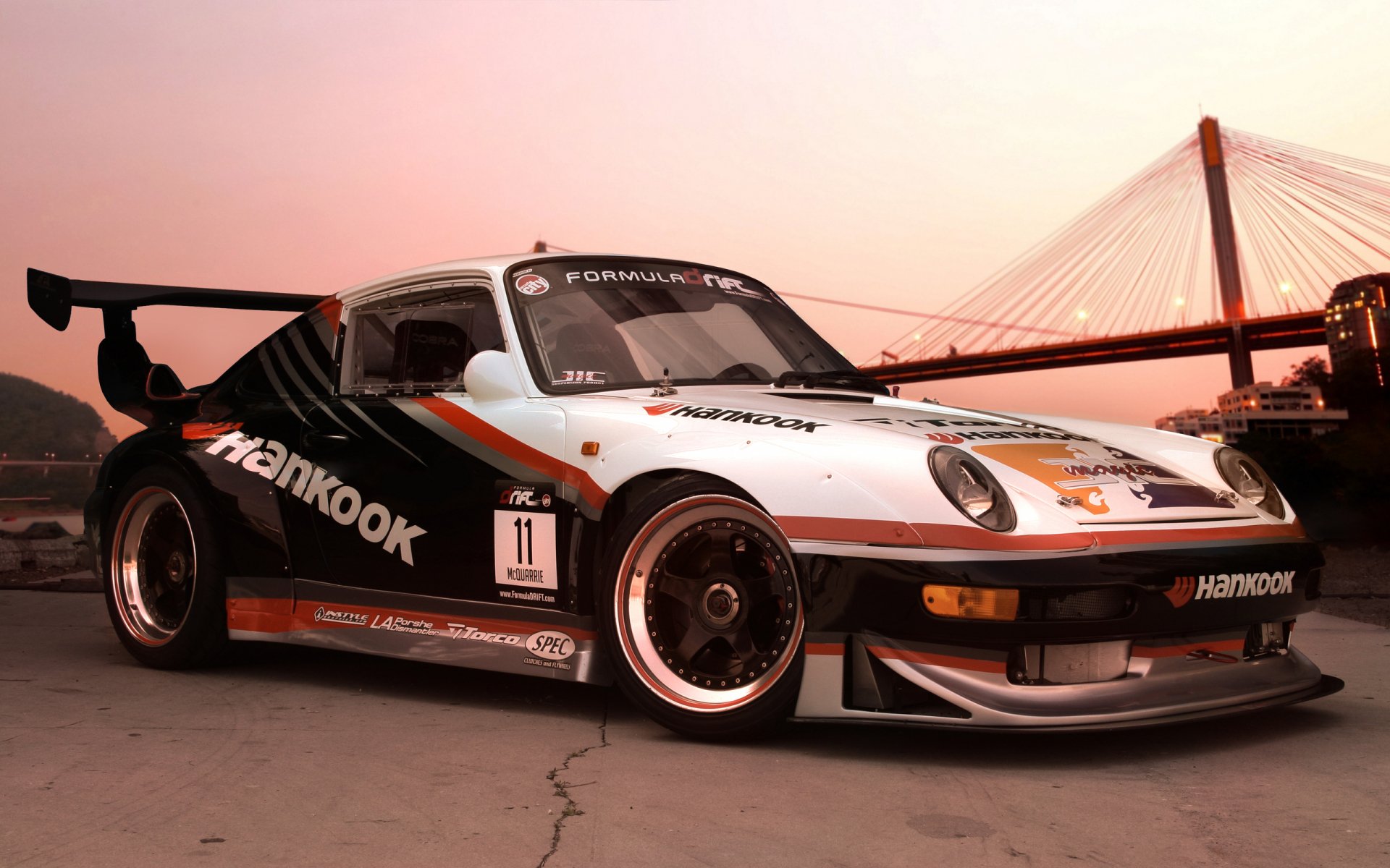 HD desktop wallpaper featuring a sleek sports car with racing decals, set against a sunset backdrop near a suspension bridge.