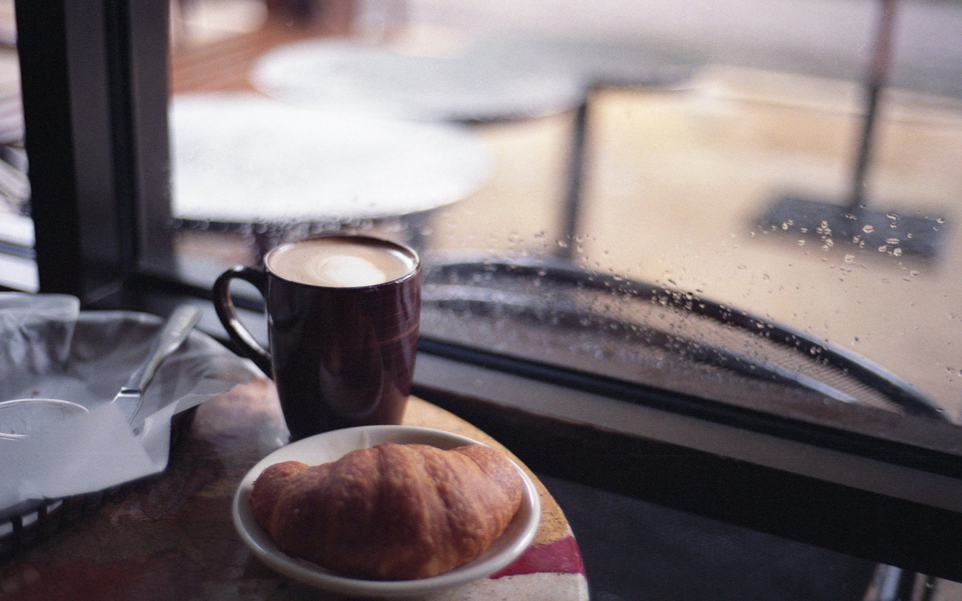 HD desktop wallpaper featuring a cozy scene of a coffee cup and a croissant on a table by a rainy window, highlighting food and coffee themes.