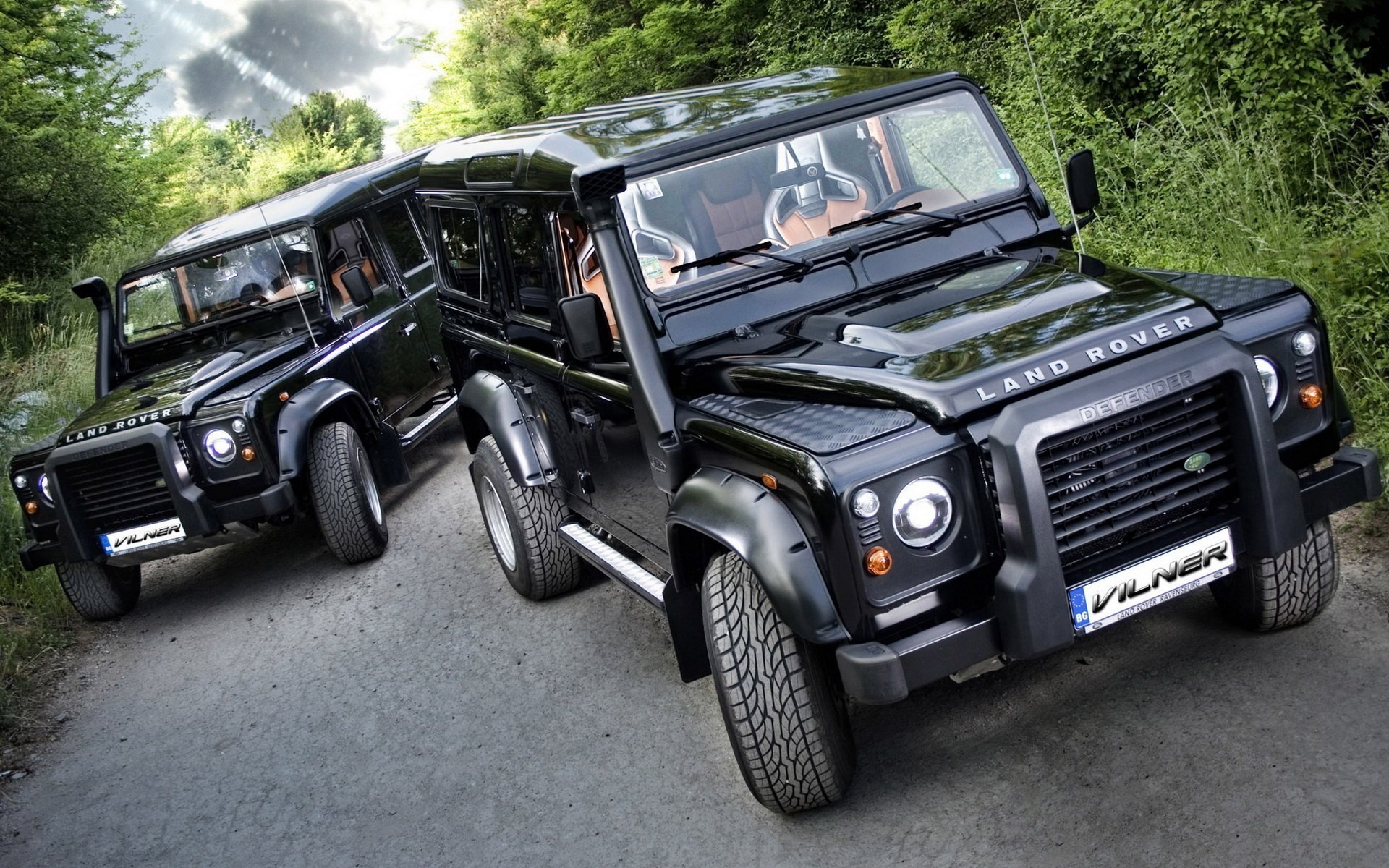 HD PC desktop wallpaper/background: two black Land Rover vehicles parked on a rural lane, glossy finish reflecting trees and sky amid lush green foliage.