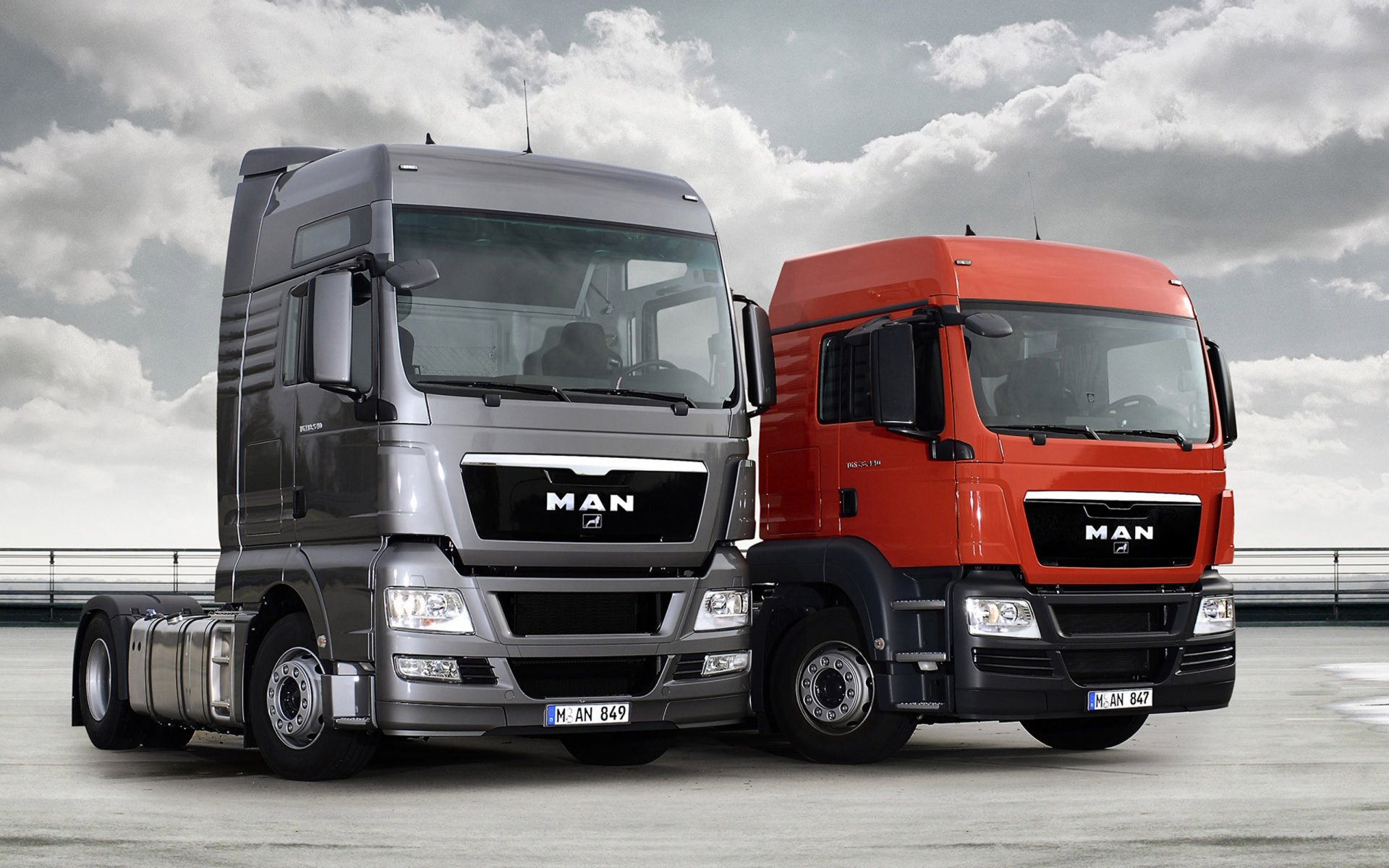 HD desktop wallpaper featuring two MAN trucks, one silver and one red, parked side by side under a cloudy sky.