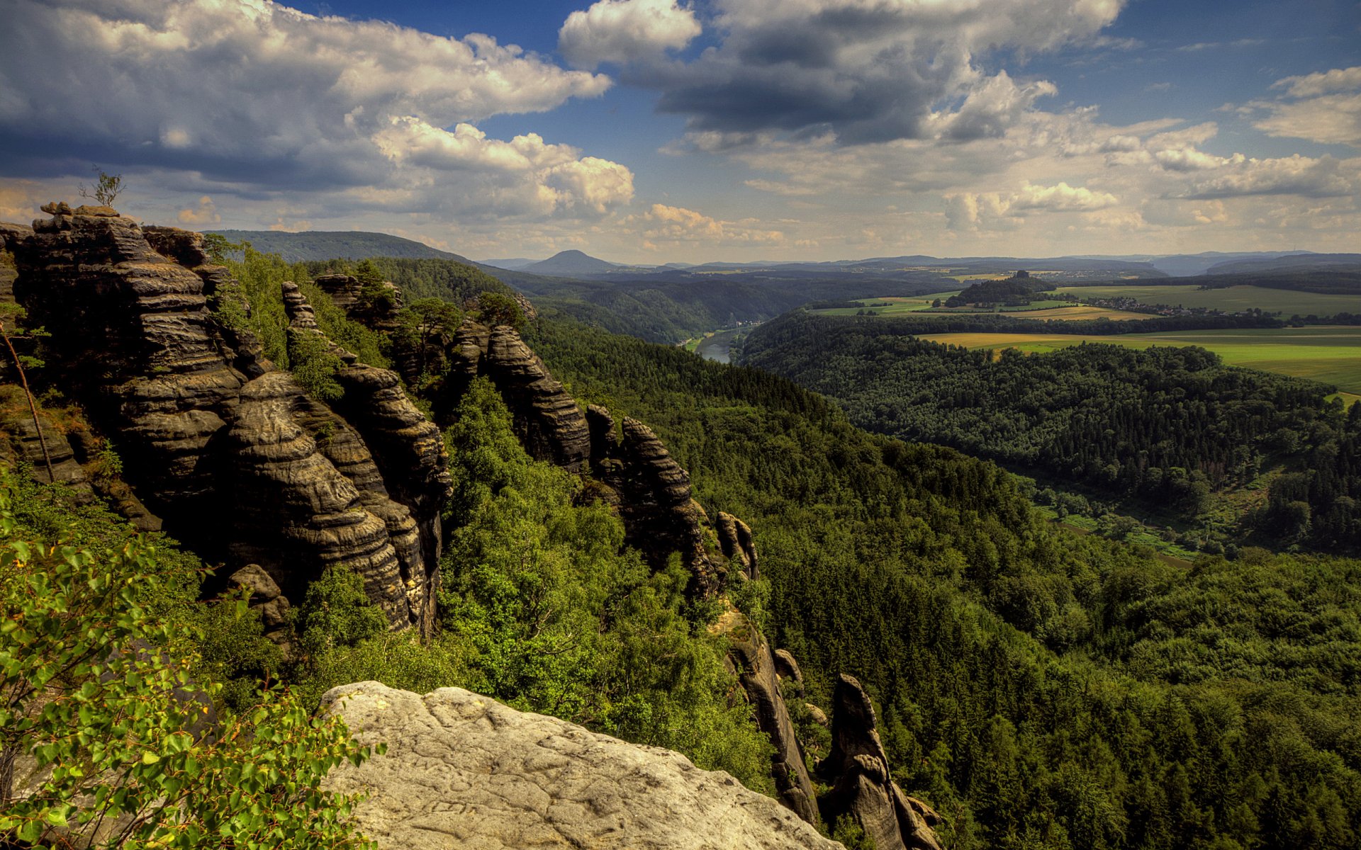 HD PC desktop wallpaper capturing a vast nature landscape with rugged rock formations, dense green forests, and a cloudy sky stretching over rolling hills.
