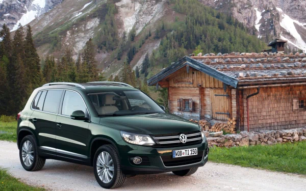 Green Volkswagen SUV parked beside a rustic mountain cabin with snow-capped peaks and pine forest — 2K Quad HD PC desktop wallpaper background.