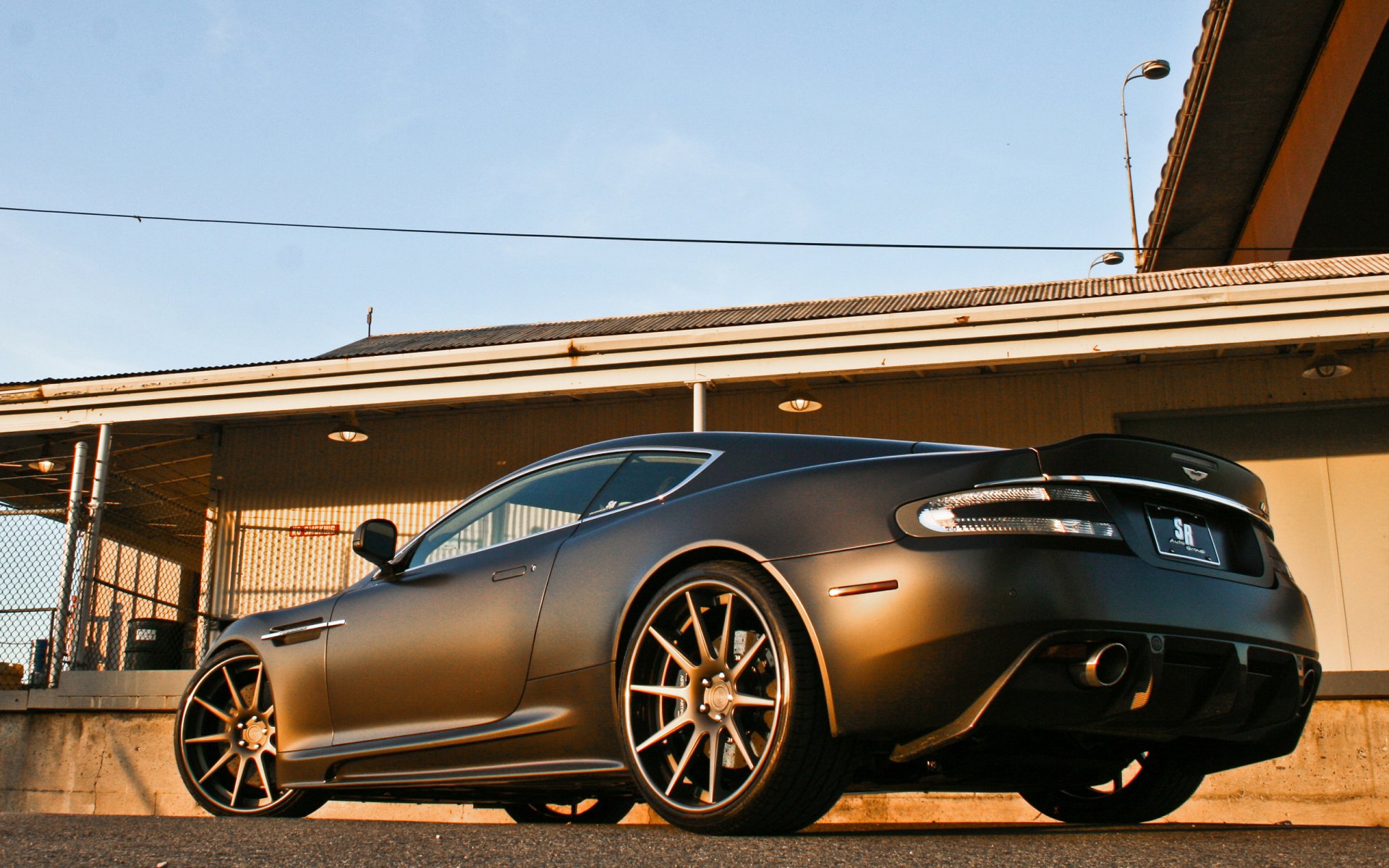 Low-angle shot of a matte-gray Aston Martin DBS parked beneath an overpass at golden hour — 2K Quad HD PC desktop wallpaper and background.