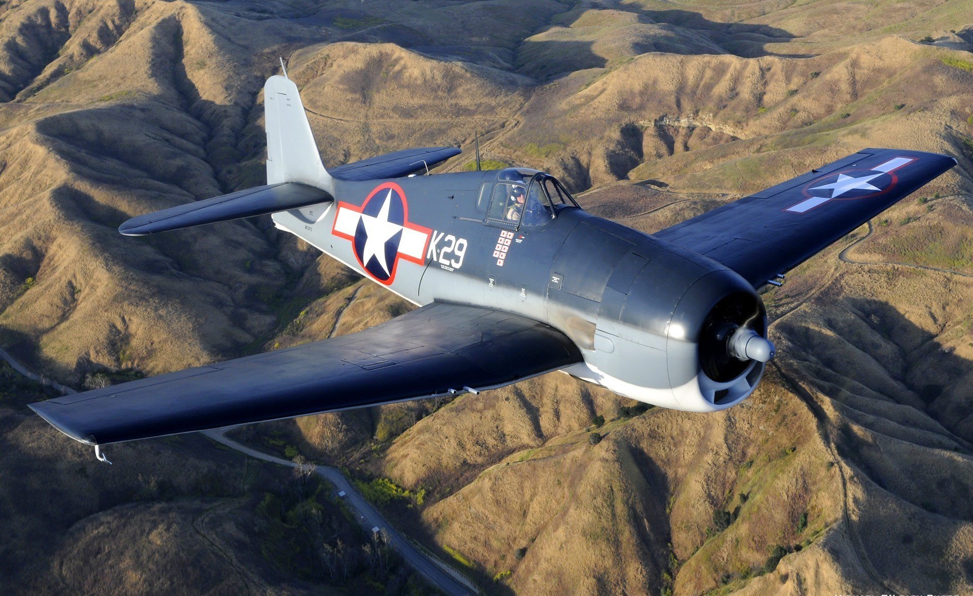 HD desktop wallpaper featuring a vintage military aircraft flying over rugged mountainous terrain, showcasing its historic design and insignia.