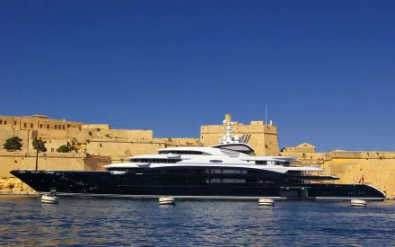 HD desktop wallpaper featuring a sleek black and white yacht docked near historic stone fortifications under a clear blue sky.