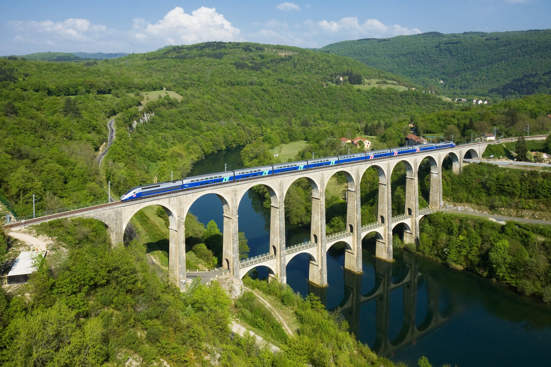 A high-definition desktop wallpaper depicting a train crossing over a scenic arched bridge surrounded by lush green hills and a river below.