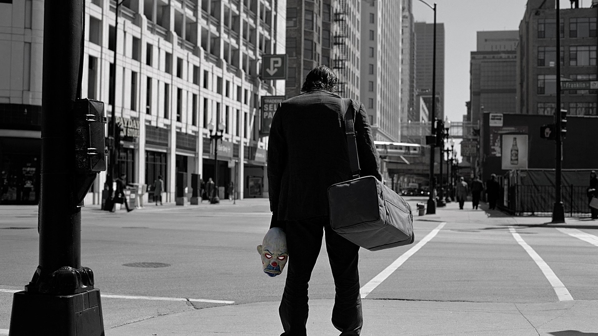 HD PC desktop wallpaper from the movie The Dark Knight, featuring a man in a suit holding a white mask, standing on a city street in a grayscale urban setting.