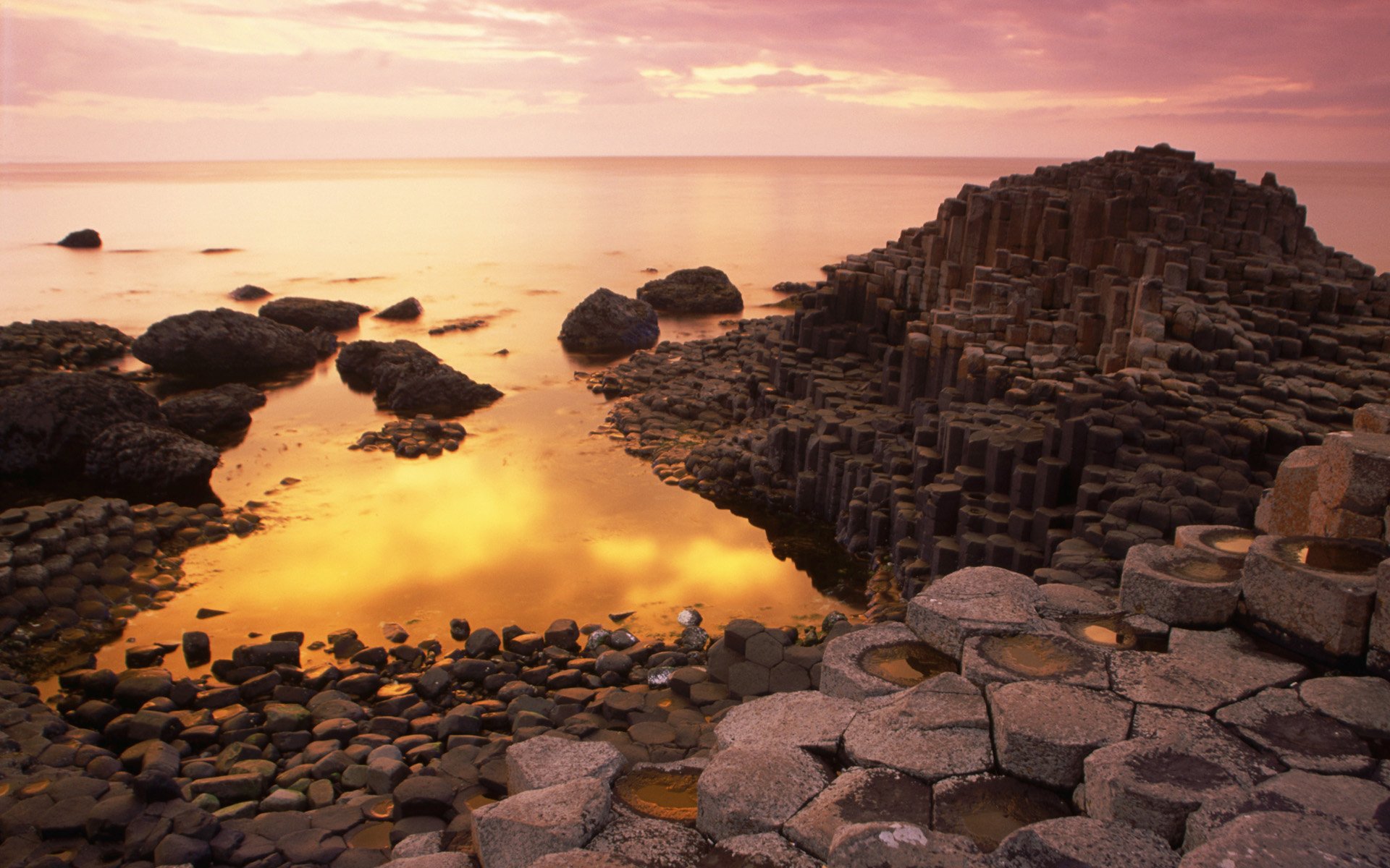 HD PC desktop wallpaper showcasing a natural rock formation with hexagonal columns by a calm body of water under a soft pink sunset sky.