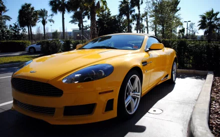 HD PC desktop wallpaper featuring a vibrant yellow Aston Martin sports car parked outdoors with palm trees and clear skies in the background.
