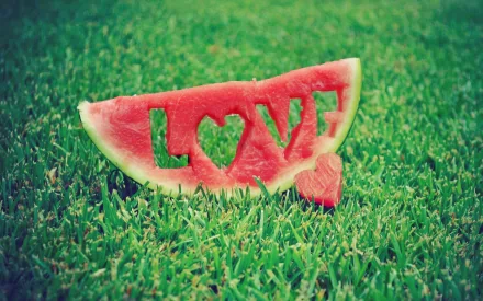 Close-up of a watermelon slice carved with the word LOVE and a heart, placed on green grass. This HD desktop wallpaper and background captures a vibrant, creative expression of love.