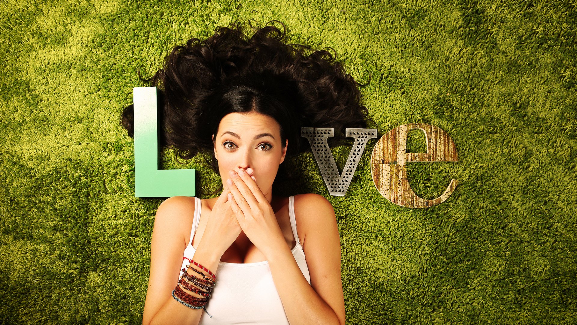 A woman with long dark hair lies on green grass, covering her mouth playfully, surrounded by artistic letters spelling Love, creating a vibrant and engaging desktop wallpaper.