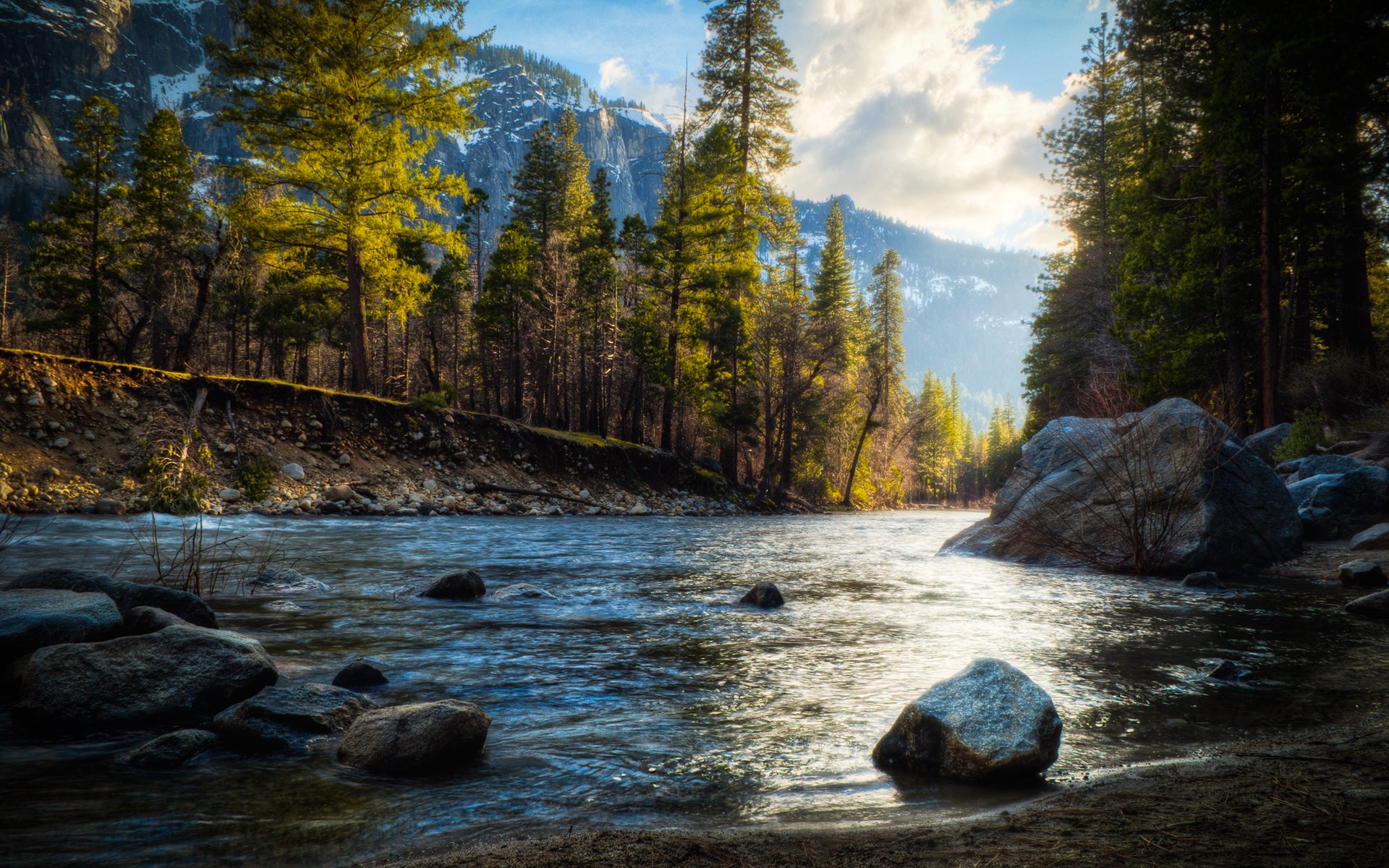 HD PC desktop wallpaper showing a peaceful river flowing through a forest with tall trees and mountains in the background under a partly cloudy sky.