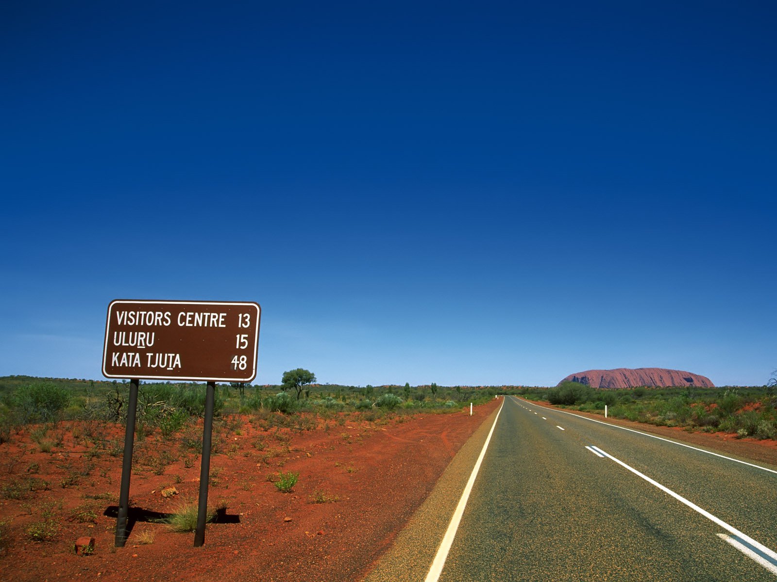 A clear blue sky stretches over a road with a directional sign indicating distances to a visitors center, Uluru, and Kata Tjuta, framed by red earth and sparse greenery.