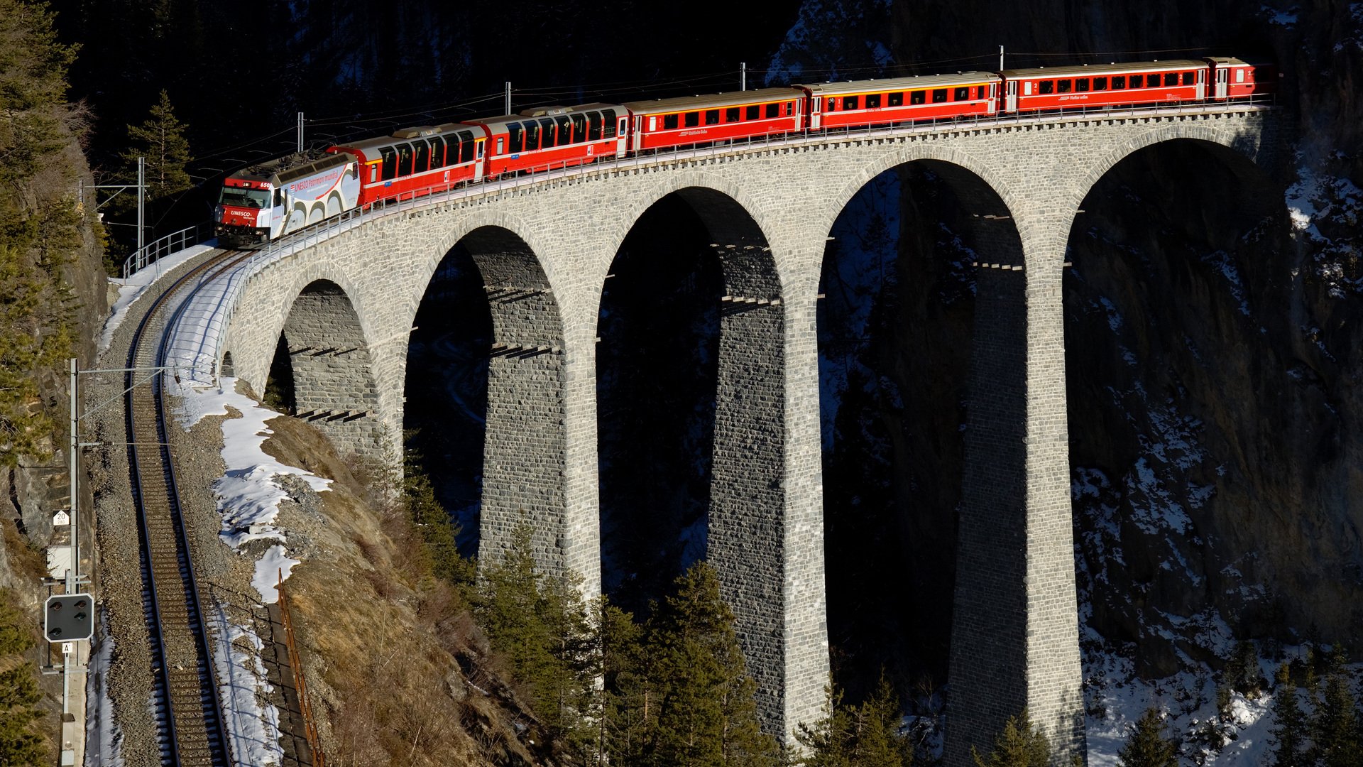 HD PC desktop wallpaper background: RhB red train (vehicle) curves across a stone viaduct above a snowy Swiss gorge.