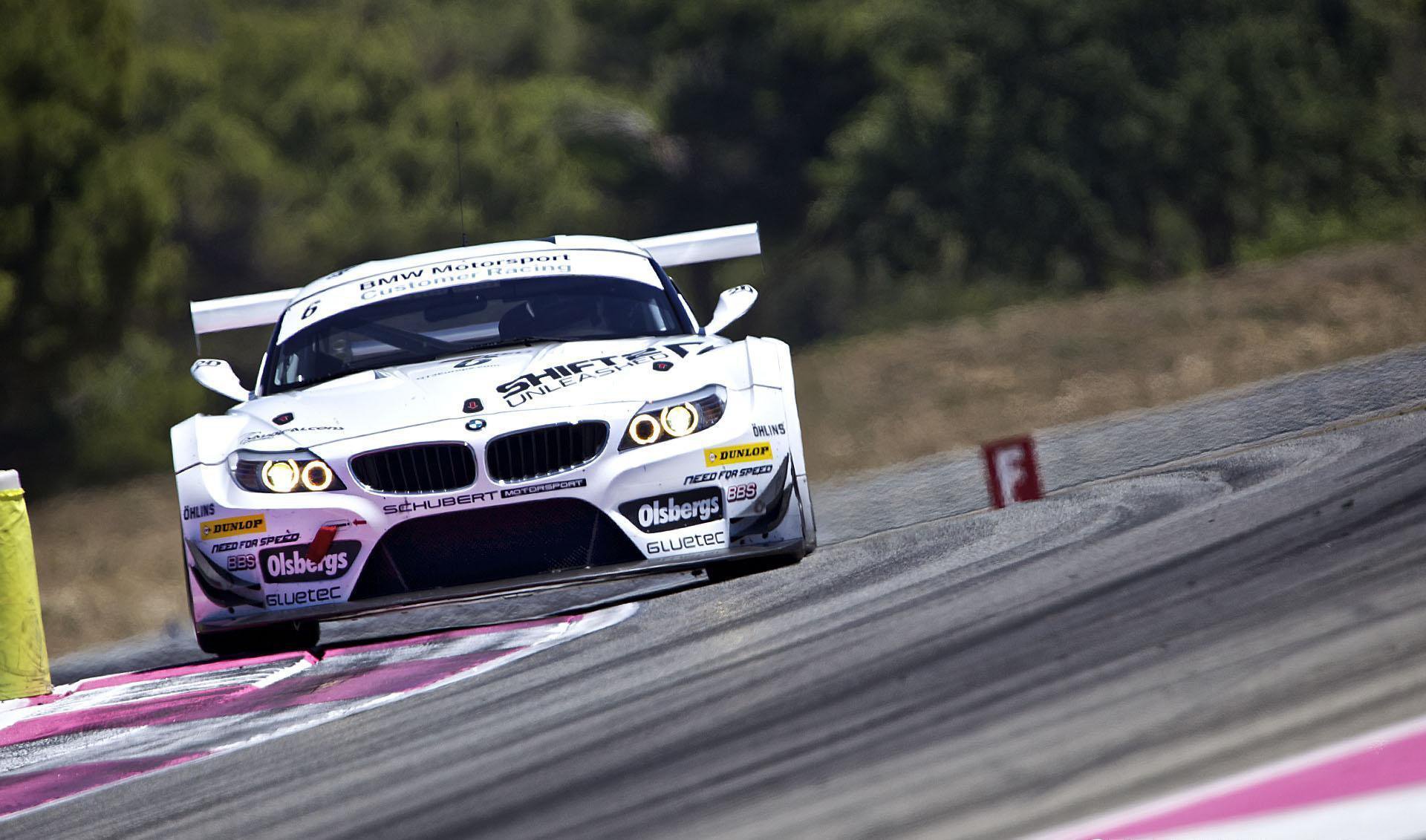 HD PC desktop wallpaper: white sports car vehicle racing through a corner on a sunlit track, front view with motion blur and trees in the background.