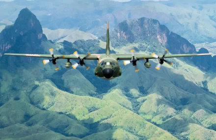 A Lockheed C-130 Hercules military aircraft soars over lush green mountains, captured in stunning detail as a high-definition desktop wallpaper and background.