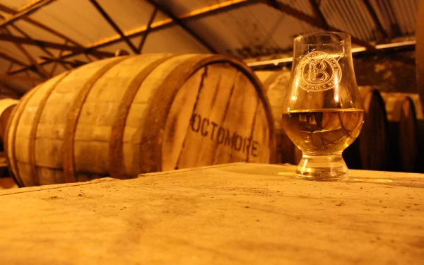 4K Ultra HD wallpaper of a whisky glass on a wooden barrel with stacked whisky casks in a warm, rustic cellar setting.