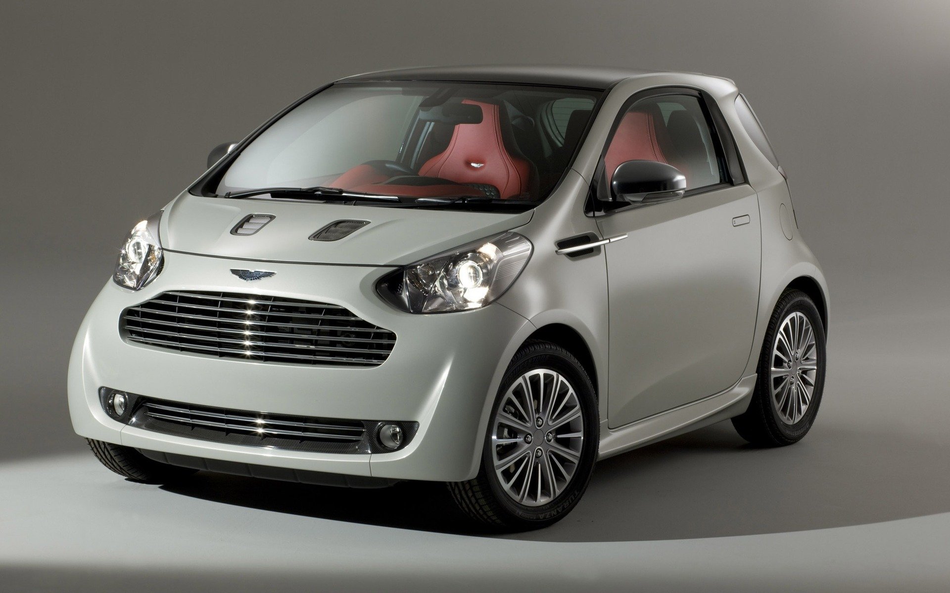 Silver 2011 Aston Martin 260 Cygnet compact car with red leather interior, studio-lit HD PC desktop wallpaper.